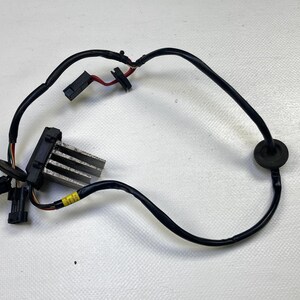 Puede incluir: Un componente eléctrico automotriz con una carcasa de plástico negro y aletas metálicas, conectado por cableado negro. El cableado incluye múltiples conectores y una arandela de goma. Se ve una etiqueta amarilla.