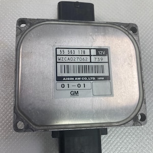 May include: A silver automotive control unit with black connectors. The unit has a label with the text "55 563 178", "WZCA027062", "12V", "739", "AISIN AW CO.,LTD. JAPAN", "01-01", and "GM".