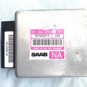 May include: A silver-coloured electronic control unit with a pink label that reads "51 64 314 BT042374 12V 248 AISIN AW CO., LTD. BELGIUM SAAB NA".