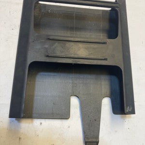 May include: Black plastic car part with a rectangular opening and a smaller rectangular opening on the side. The part has a raised lip around the edges.
