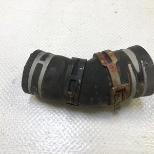 May include: A black, curved rubber hose with metal clamps. The hose has two silver bands and is secured by three clamps, one of which shows signs of rust. The hose is likely an automotive or industrial part.