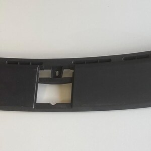 May include: A black, curved automotive dashboard component. The piece has a rectangular central section with a square opening and a series of small rectangular vents along the top edge. The ends have a flared design.