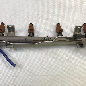 May include: A metallic fuel rail assembly featuring four brown fuel injectors mounted in a row. The component includes a silver-toned metal rail, integrated mounting brackets, a thin silver fuel line, and a short blue rubber hose attached to one end.