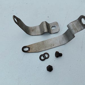 May include: Two curved, silver-colored metal mounting brackets with circular holes at each end, accompanied by two small metal washers, one bolt with a hexagonal head, and one additional hexagonal nut, all displayed on a plain light surface.