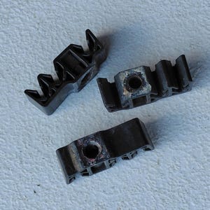 May include: Three black plastic mounting brackets, each featuring a central circular hole and a series of parallel ridges along one side. These components are designed for securing items and have a rigid, molded construction with a matte finish.