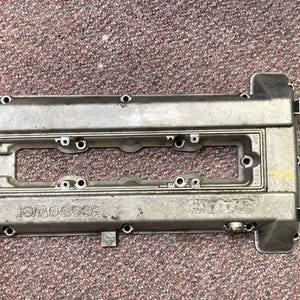 May include: A silver-colored engine valve cover with multiple bolt holes and the word "SAAB" and "ecopower" embossed on the surface. The cover has a rectangular shape with a central cutout.