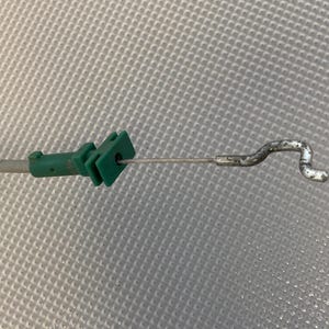 Puede incluir: Una pieza de automóvil de plástico verde con un cable metálico y un extremo curvado. La pieza tiene forma rectangular con un agujero para el paso del cable. El cable es de color plateado.