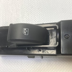 May include: A black car window switch with a rocker button featuring a window icon with arrows. The switch is part of a larger black plastic assembly, likely for a vehicle's interior controls.