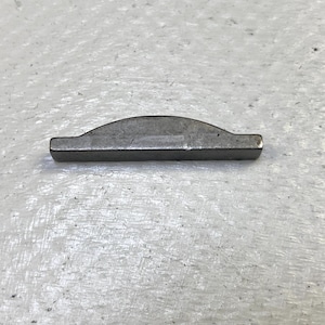 May include: A small, silver-colored metal key, with a curved top and flat bottom. The key is rectangular with rounded edges and is likely used for machinery or mechanical applications. The key is set against a light gray, textured surface.