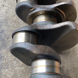 May include: Close-up of a used, dark gray crankshaft. The crankshaft has multiple cylindrical bearing surfaces with silver-colored metal rings. The metal has a slightly oily appearance. The background is a light gray concrete surface.