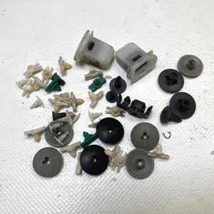 May include: A collection of various automotive plastic fasteners and clips in shades of black, gray, green, and off-white. The assortment includes push-type retainers, panel clips, and other small hardware components, scattered on a white surface.