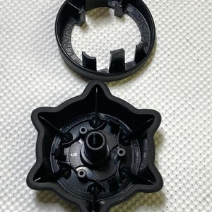 May include: Two black plastic components, one circular with a notched edge and the other star-shaped with a central hole and several smaller holes. The parts are on a white, textured surface.
