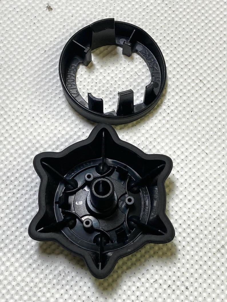 May include: Two black plastic components, one circular with a notched edge and the other star-shaped with a central hole and several smaller holes. The parts are on a white, textured surface.