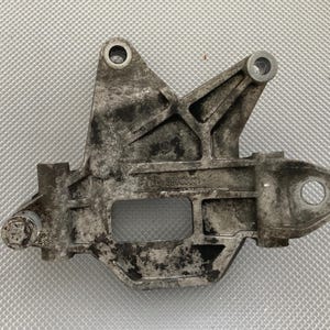 May include: A weathered, silver-colored metal bracket with multiple holes and a rectangular cutout. The bracket has a complex shape with angled supports and appears to be a component of a larger mechanical assembly. The surface shows signs of wear.