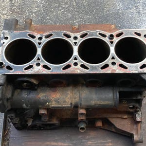 May include: A used engine block with four large cylindrical openings. The metal component shows signs of wear, with rust and discoloration. The top surface has a silver border with various openings and a red-brown gasket.