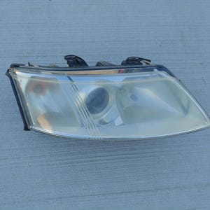 May include: A vehicle headlight assembly featuring a clear, slightly yellowed polycarbonate lens, a central circular projector lens, and a black plastic housing with mounting brackets visible along the top edge of the unit.