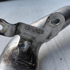 May include: A metallic automotive pipe component featuring a mounting bracket with two circular holes. The bracket is stamped with the text SAAB, the number 12785078, and the number 50029. The metal surface shows signs of wear and dark residue.