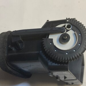以下が含まれることがあります： 黒いプラスチック製のギア機構で、白いギアと黒いレバーが付いています。ギア機構は黒い布製のストラップに取り付けられています。