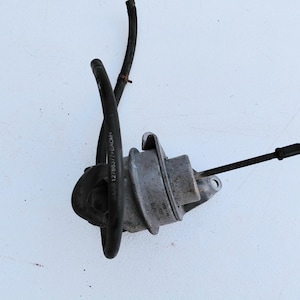 May include: A metallic automotive wastegate actuator featuring a circular housing, a protruding metal rod with a circular end, and two attached black rubber hoses. The housing displays printed text including SAAB 127865377 and other markings.