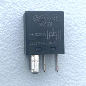 May include: A black rectangular automotive relay featuring the SAAB logo and the part number 5604301 printed in white. The component displays a 12V rating, a circuit diagram, and the identification code V23074-A2501-X78 with three metal terminals.