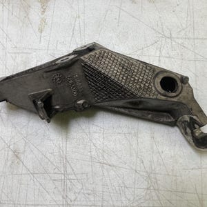 May include: A silver-colored metal automotive part with a textured surface and multiple holes. The part has a triangular shape with a curved edge and a small, rectangular extension. The part appears to be used in a vehicle's engine or chassis.