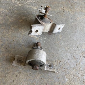May include: Two used, silver-colored automotive engine mounts. Each mount has a rubber bushing, a metal bracket with bolt holes, and a threaded stud with a nut. The mounts are on a gray concrete surface.