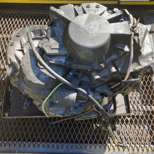 May include: A close-up of a gray metal automotive transmission with black hoses and wires attached. The transmission is sitting on a metal grate.