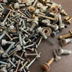 May include: A large collection of assorted screws, bolts, and nuts in various sizes and finishes. The metal hardware is scattered on a wooden surface.