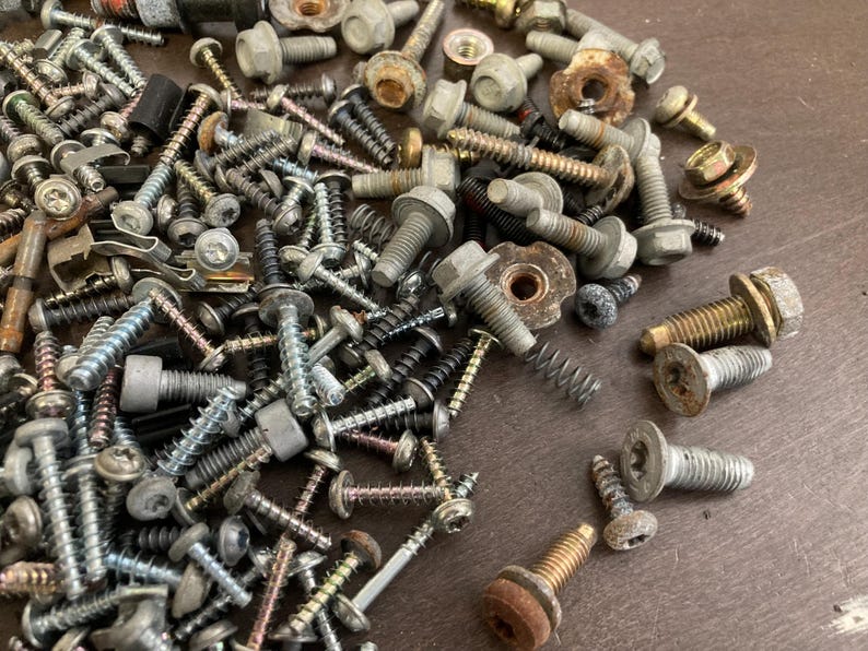 May include: A large collection of assorted screws, bolts, and nuts in various sizes and finishes. The metal hardware is scattered on a wooden surface.