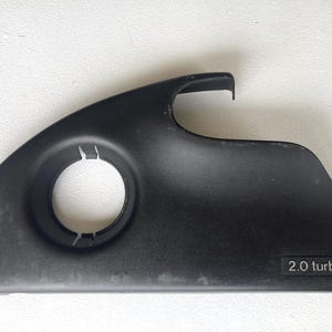 May include: A black plastic automotive engine cover component featuring a circular opening on the left side and a curved top edge. The lower right corner displays the embossed text 2.0 turbo in a rectangular frame, indicating engine specifications.