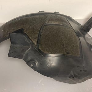 Puede incluir: Revestimiento de guardabarros de coche de plástico negro con un material texturizado y amortiguador de sonido. El revestimiento tiene una pequeña pestaña elevada en el lateral.