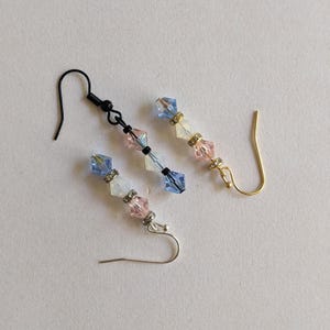 May include: Three pairs of dangle earrings with colorful beads. Each earring features a series of blue, pink, and white faceted beads, separated by silver-colored spacers. The earring hooks are black, gold, and silver.