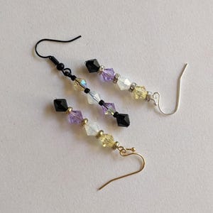 May include: Three pairs of beaded earrings with black, purple, white, and yellow beads. The earrings have different colored hooks: black, silver, and gold. The earrings are arranged on a white surface.