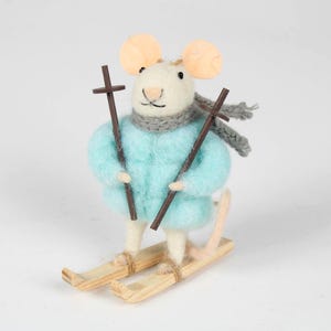 May include: A whimsical felt mouse figurine dressed for skiing. The mouse wears a light blue jacket, gray scarf, and holds brown ski poles. It stands on wooden skis, with large, tan ears and a pink tail. A charming winter-themed decoration.