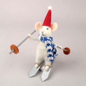 May include: A white felt mouse figurine wearing a red hat with a white pom-pom, a blue and white scarf, and skis. The mouse holds ski poles with brown circular ends. A whimsical winter-themed decoration.
