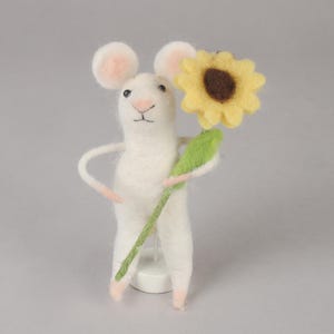 May include: A white felt mouse holding a yellow felt sunflower with a brown center. The mouse is standing on a white base.