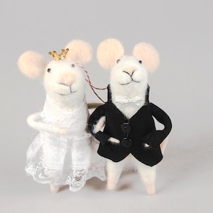 May include: Two white felt mice dressed for a wedding. One mouse wears a white lace wedding dress and a gold crown. The other mouse is in a black tuxedo with a white bow tie. The mice have pink ears and noses.