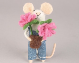 May include: A handcrafted felt mouse figurine wearing denim overalls, holding a brown pot with two pink flowers. The mouse has cream-colored ears and a long, thin tail. The flowers have green stems and leaves.