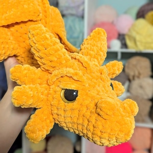 May include: A crocheted orange dragon plush toy with black eyes. The dragon has a long snout and a spiky back.