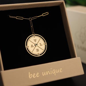 May include: Gold-colored pendant necklace in an open gift box. The pendant is circular with the letters V, A, L, and B, and a small green gem. The box is beige with the words "bee unique" printed on the front.