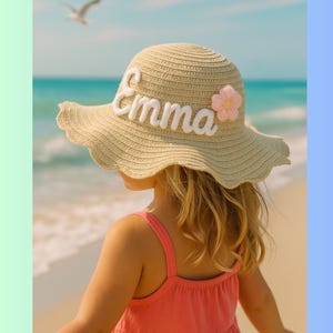 May include: A beige sun hat with a wide, wavy brim. The name "Emma" is embroidered in white script across the front, with a pink flower accent. The hat is worn by a child on a beach, with the ocean in the background.