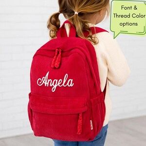 May include: A red corduroy backpack embroidered with the name "Angela" in white script. The backpack features a front pocket, side pocket, and a top handle. A green speech bubble displays "Font & Thread Color options".