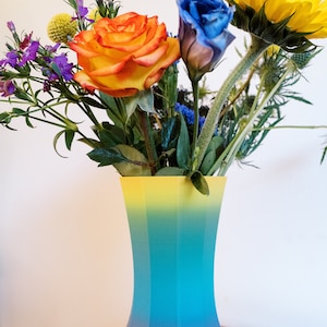 May include: A rainbow ombre vase with a yellow top and pink bottom holds a bouquet of flowers including a yellow sunflower, a blue rose, an orange rose, and purple wildflowers.