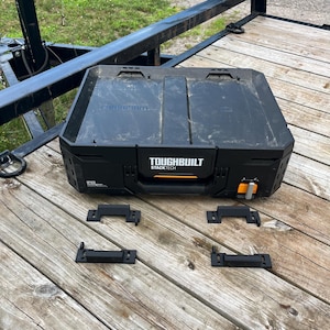 May include: Black TOUGHBUILT STACKTECH tool box with orange accents and four black brackets on a wooden surface. The tool box has a handle and the text "TOUGHBUILT STACKTECH" is visible.