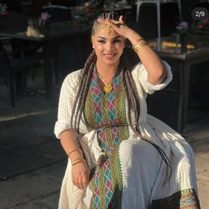 Puede incluir: Una mujer con un vestido blanco tradicional con bordados coloridos en el corpiño y las mangas. Tiene el pelo largo trenzado, joyas de oro y una pieza de cabeza dorada. El vestido presenta un patrón detallado de diseños geométricos rojos, verdes y amarillos.