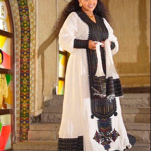 Puede incluir: Una mujer vistiendo un vestido tradicional etíope blanco con bordados negros y dorados. El vestido tiene una falda larga y mangas largas. Está de pie en un tramo de escaleras.