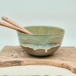 Könnte beinhalten: Eine runde Keramikschale mit mattbrauner Basis und einer glänzenden grünen Glasur, die an den Seiten herabläuft. Zwei helle hölzerne Salatbestecke befinden sich in der Schale. Ein kleiner Zweig getrockneter weißer Blumen liegt neben der Schale.