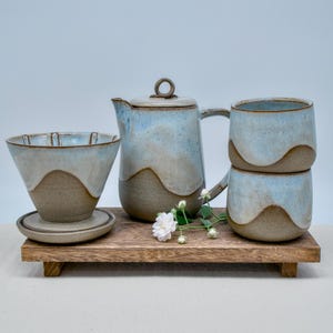 May include: A ceramic coffee set featuring a pour-over dripper with a matching saucer, a lidded pitcher, and two stacked mugs. Each piece has a two-tone design with a textured brown base and a smooth, glossy light blue glaze on the upper portion.