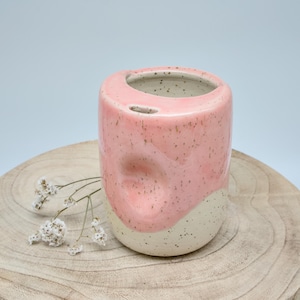 Könnte beinhalten: Ein zylindrischer Keramikbecher mit einer gesprenkelten cremefarbenen Basis und einem glänzenden rosa Oberteil. Der Becher hat eine deutliche Vertiefung an einer Seite und eine kleine Einkerbung am Rand. Ein Zweig mit kleinen weißen Blüten liegt daneben.