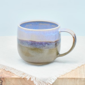 Könnte beinhalten: Eine Keramik-Tasse mit rundem Körper und Henkel. Die Tasse hat einen Farbverlauf, beginnend mit einem blauen Rand, übergehend in Lavendel, dann ein lilafarbenes Band und endend mit einem grün-braunen Boden. Die Tasse steht auf einem weißen Tuch.